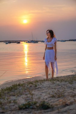 Güzel bir model gün batımında beyaz bir elbise giyerek kıyıda duruyor. Sakin su, batan güneşin sıcak tonlarını yansıtarak, İspanya 'nın Balear Adaları, Formentera' da sakin bir atmosfer oluşturur..