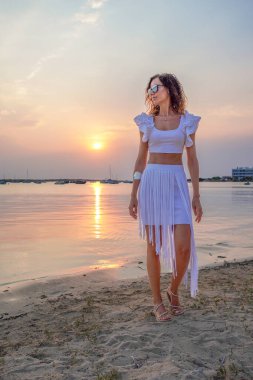 Günbatımında sahilde gezen bir kadın, Formentera, Balearic Adaları, İspanya 'daki kıyı şeridinin sakin atmosferini tamamlayan şık bir yaz kıyafeti giyer..