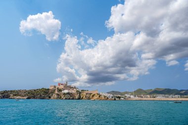 Kayalık bir sahil şeridinde, berrak mavi sular ve tüylü beyaz bulutlarla çelişen tarihi bir kalenin yer aldığı nefes kesici bir sahil manzarası. Eivissa, İbiza, İspanya.