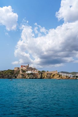 Parlak mavi suları ve canlı bulutlarıyla kayalıklara tünemiş tarihi bir şatonun bulunduğu çarpıcı deniz manzarası. Eivissa, İbiza, İspanya.