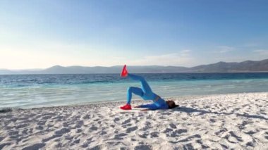 Kadın, deniz ya da okyanus manzaralı bir plajda yoga yaparken görülüyor. Mavi spor takım elbiseli kadın bir dizi yoga pozunda zarafet ve akışkanlıkla hareket ediyor. Kumsal.