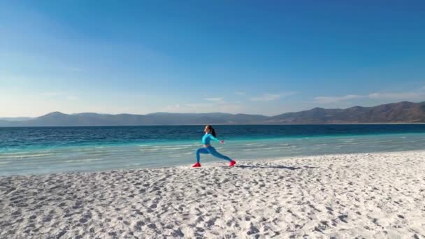 海や海の素晴らしい景色を望むビーチでヨガを練習している女性が表示されます 青いスポーツスーツを着た女性は 優雅さと流動性を持つヨガのポーズのシリーズを通って移動します ビーチ — ストック動画