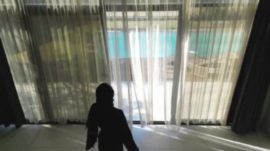 Siyah abaya giymiş bir Arap kadının yüzme havuzlu bir villanın perdelerini açtığı görülüyor. Geleneksel kadın kıyafetleri ve lüks villa manzarası...