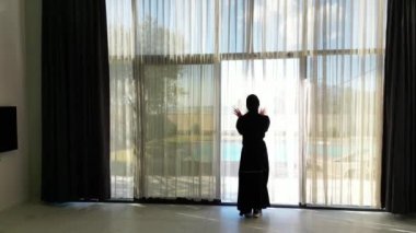 Siyah abaya giymiş bir Arap kadının yüzme havuzlu bir villanın perdelerini açtığı görülüyor. Geleneksel kadın kıyafetleri ve lüks villa manzarası...