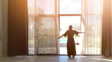 Siyah abaya giymiş bir Arap kadının yüzme havuzlu bir villanın perdelerini açtığı görülüyor. Geleneksel kadın kıyafetleri ve lüks villa manzarası...