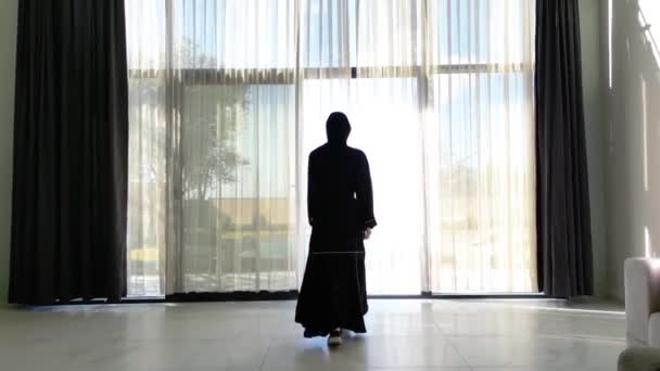 黒いアバヤに身を包んだアラブ人女性がプール付きの別荘のカーテンを開けているのが見える 女性の伝統的な服やヴィラの豪華な設定は コントラストを提供します — ストック動画