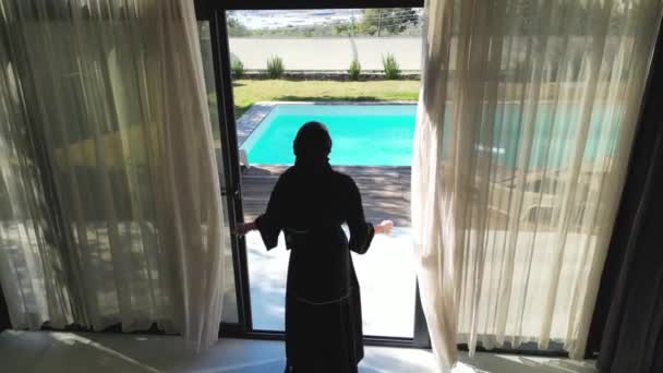 黒いアバヤに身を包んだアラブ人女性がプール付きの別荘のカーテンを開けているのが見える 女性の伝統的な服やヴィラの豪華な設定は コントラストを提供します — ストック動画