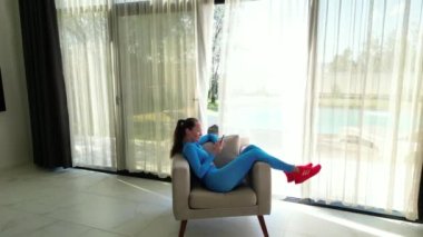 The video features a woman sitting comfortably in a chair near a large window in a luxurious villa. She is dressed in a blue sport suit, looking relaxed and focused as she uses her smartphone. The
