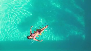 In this video, we see an aerial shot of a pool. The camera is high up, and we can see the crystal clear water and sun loungers around the edge. The pool is surrounded by lush greenery, providing a