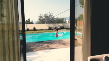 In this video, we see a camera moving through a window, showing a luxurious swimming pool, surrounded by lush greenery and beautiful architecture. The pool is crystal clear and inviting, with sun