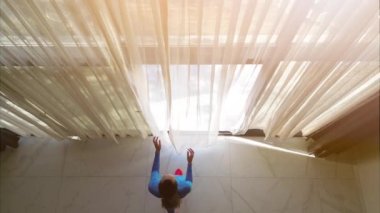 In this video, we see a young woman, dressed in a stylish blue sport suit, walking towards a large set of curtains in a luxurious villa. With a flick of her wrist, she opens the curtains to reveal a