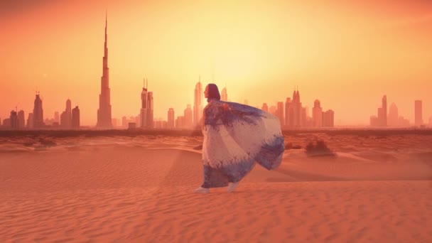 広大な砂漠の風景の中に立つ伝統的なアラブの服を着た女性の美しいシーンを撮影しています 背景にはドバイの美しいスカイラインがシルエットになっています — ストック動画