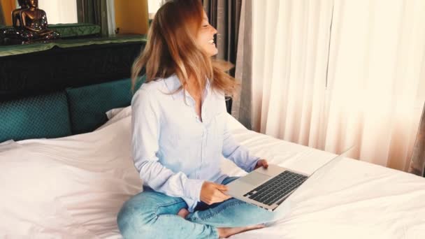 快乐漂亮的女性从家里工作在关键笔记本电脑从卧室 慢动作 使用笔记本电脑应用程序的年轻漂亮的女人穿着睡衣坐在舒适的床上 高质量的4K镜头 — 图库视频影像