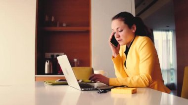 Sarı tişörtlü, kulaklık takan, mutfaktaki dizüstü bilgisayarda çalışan ve masada yeşil meyve püresi olan genç, sarışın bir kadın. Yüksek kalite 4k görüntü