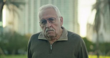 Medium shot of a senior man looking at the camera with a serious expression while at a park