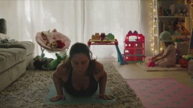 Annesi evde yoga yaparken küçük kızı arka planda oyuncaklarla oynuyor.