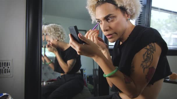 小さな鏡を押しながら大きな鏡の隣に座ってメイクを入れてトランスジェンダーの女性のスローモーション撮影 — ストック動画