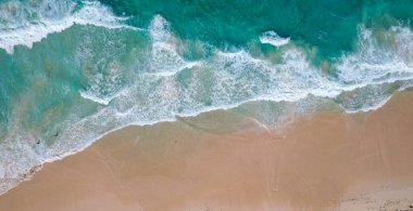 Sahile çarpan turkuaz dalgaların olduğu kumlu bir sahilin havadan görünüşü sahil boyunca yürüyen dağınık insanlar görülüyor..