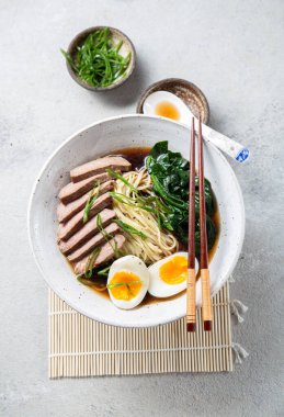 Bir kase bonfile, kaynamış yumurta ve yeşil sebzeli ramen. Yemek yeşil soğanla süsleniyor ve yanında yemek çubukları ve örülmüş bir kaşık var.