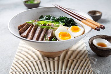 Bir kase bonfile, kaynamış yumurta ve yeşil sebzeli ramen. Yemek yeşil soğanla süsleniyor ve yanında yemek çubukları ve örülmüş bir kaşık var.