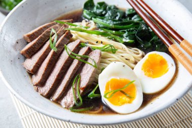 Bir kase bonfile, kaynamış yumurta ve yeşil sebzeli ramen. Yemek yeşil soğanla süsleniyor ve yanında yemek çubukları ve örülmüş bir kaşık var.