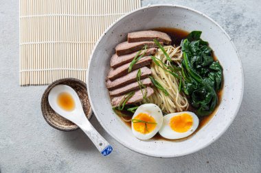 Bir kase bonfile, kaynamış yumurta ve yeşil sebzeli ramen. Yemek yeşil soğanla süsleniyor ve yanında yemek çubukları ve örülmüş bir kaşık var.