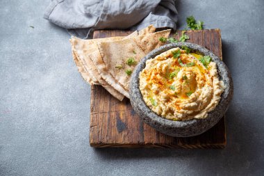 Geleneksel Yahudi humusu, zeytinyağı, paprika ve maydanozla süslenmiş taştan bir kasede servis edilir. Yanında da flatekmek ve gri bir masada taze otlar bulunur..