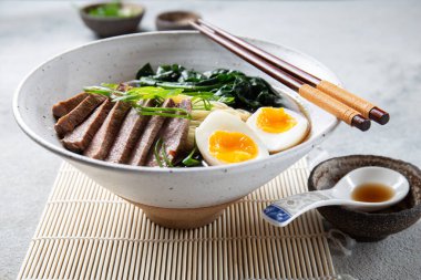 Bir kase bonfile, kaynamış yumurta ve yeşil sebzeli ramen. Yemek yeşil soğanla süsleniyor ve yanında yemek çubukları ve örülmüş bir kaşık var.