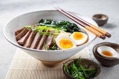 Bir kase bonfile, kaynamış yumurta ve yeşil sebzeli ramen. Yemek yeşil soğanla süsleniyor ve yanında yemek çubukları ve örülmüş bir kaşık var.