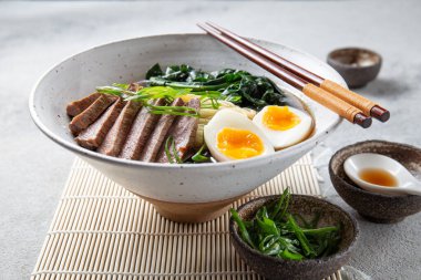 Bir kase bonfile, kaynamış yumurta ve yeşil sebzeli ramen. Yemek yeşil soğanla süsleniyor ve yanında yemek çubukları ve örülmüş bir kaşık var.