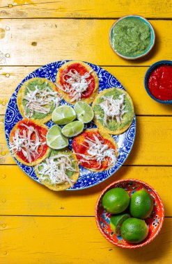 Parçalanmış tavukla kaplı bardaklar ve canlı yeşil ve kırmızı soslar renkli bir tabakta düzenlenir. Limonlar ve salsa kaseleri sarı masayı süslüyor ve festival sunumunu güçlendiriyor..