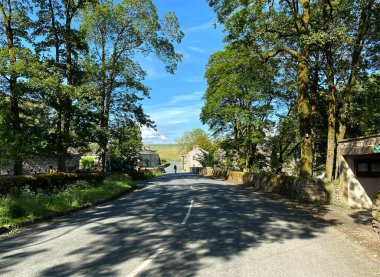 Linton, Skipton, İngiltere 'deki Yorkshire Dales köyünde, eski ağaçlar, evler ve uzak tepeler olan bir yol manzarası.