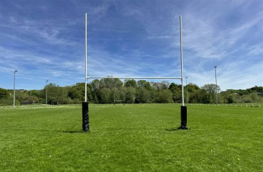Bir çift rugby kale direği mavi gökyüzünün altındaki yemyeşil bir sahanın ortasında belirgin bir şekilde duruyor. Arka planda, Cottingley, İngiltere 'deki oyun alanının kenarı ağaçlarla çevrili.