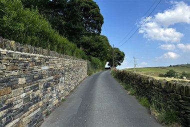Kırsal bir alanda dar bir yol esiyor, her iki tarafında kuru taş duvarlar ve yemyeşil alanlar var. Gökyüzü açık ve mavi. Brow Lane, Clayton, İngiltere 'de birkaç dağınık bulut var.