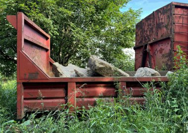 Büyük kayalarla dolu kırmızı metal bir konteynır, ağaçlarla benekli yemyeşil bir çimenliğin yanında yuva yapar. Yakınlarda, başka bir kırmızı konteynır Wrose, Bradford, İngiltere 'de zamanın işaretlerini taşıyor.