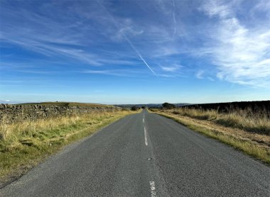 Uzun, boş bir Yeşil Sykes yolu mavi gökyüzünün altında uzanır. Çimenli tarlalar ve her iki tarafında da taş duvarlar vardır. Gökyüzü Oakworth, İngiltere yakınlarında ince bulutlarla süslenmiş.
