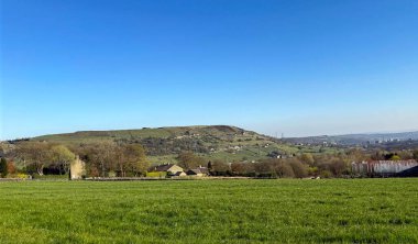 Masmavi bir gökyüzünün altında yemyeşil bir alan uzanır. Arkasında yuvarlanan tepeler ve dağınık konutlar vardır. Uzak yapılar ve ağaçlar uyum içinde, Catherine Slack Queensbury, İngiltere