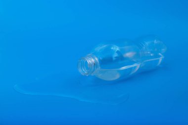 Mavi zemin üzerine dökülen suyla yan yatmış plastik bir su şişesinin yakın plan çekimi, kaza konsepti, atık, su ve tazelikler