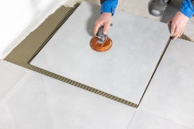 Tiler worker placing or tiling gray ceramic tile in position over adhesive glue with lash tile leveling system, renovation or recontruction, concept of building