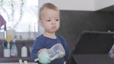 Küçük çocuk tabletten bir şey izlerken eğitim bardağından içer.