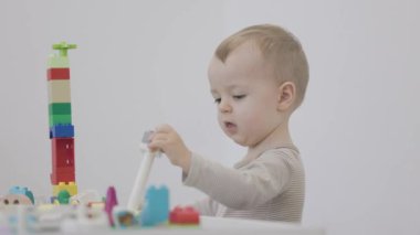 Bebek bir masada oturuyor ve beyaz arka planda renkli yapı taşlarıyla oynuyor.