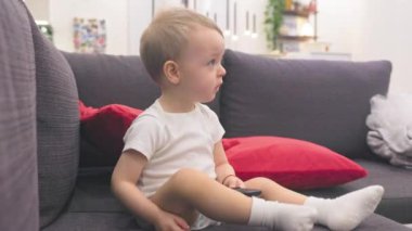 Sevimli bebek kanepede oturup televizyon izliyor ve uzaktan kumandayla kanal değiştiriyor.