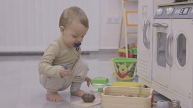 Oyuncak mutfağın yanında tahta yemek ve emzik ile oynayan bir bebek.