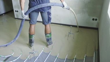 Profesyonel inşaat işçisi ustalıkla yeraltı ısıtma borularının üzerine sıvı çimento dökerek zemini döşemek için pürüzsüz ve eşit bir yüzey oluşturuyor.