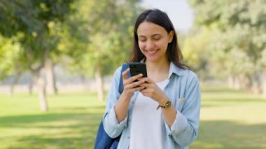 Happy Indian girl typing and chatting on mobile phone at a park
