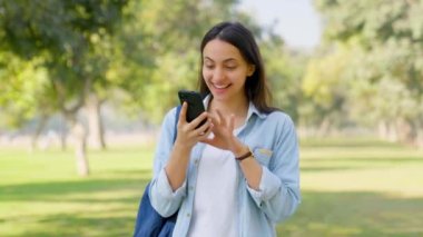 Happy Indian girl scrolling through phone in a park in sunlight