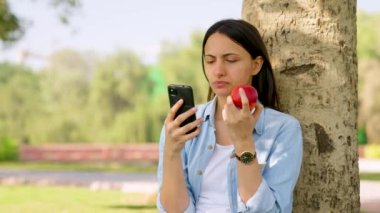 Indian girl eating an apple while using her phone for breakfast in a park
