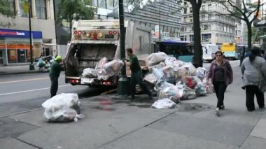 Garbage Truck in NYC and two DSNY workers Loading White Plastic Garbage Trash Bags into Truck. People on Sidewalk. Manhattan. Morning.