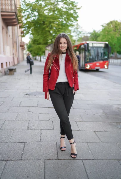 美丽的少女站在立陶宛维尔纽斯老城的人行道上 穿着红色夹克和黑色裤子 美丽的春天 笑个不停背景下的公共交通巴士 — 图库照片