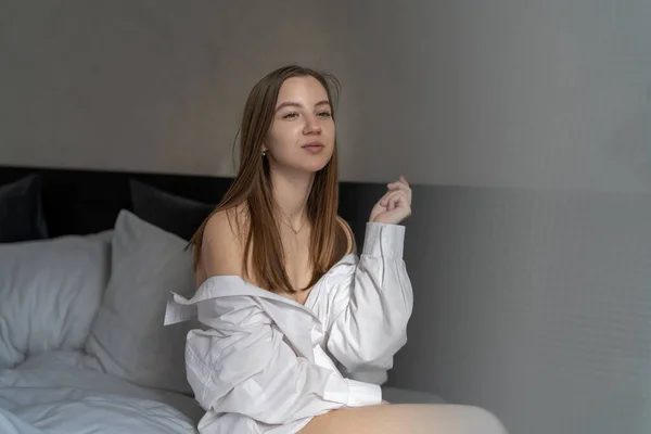 美しい若い女の子はベッドルームでベッドに座っています 朝の日光と影の体に 白いシャツを着て — ストック写真